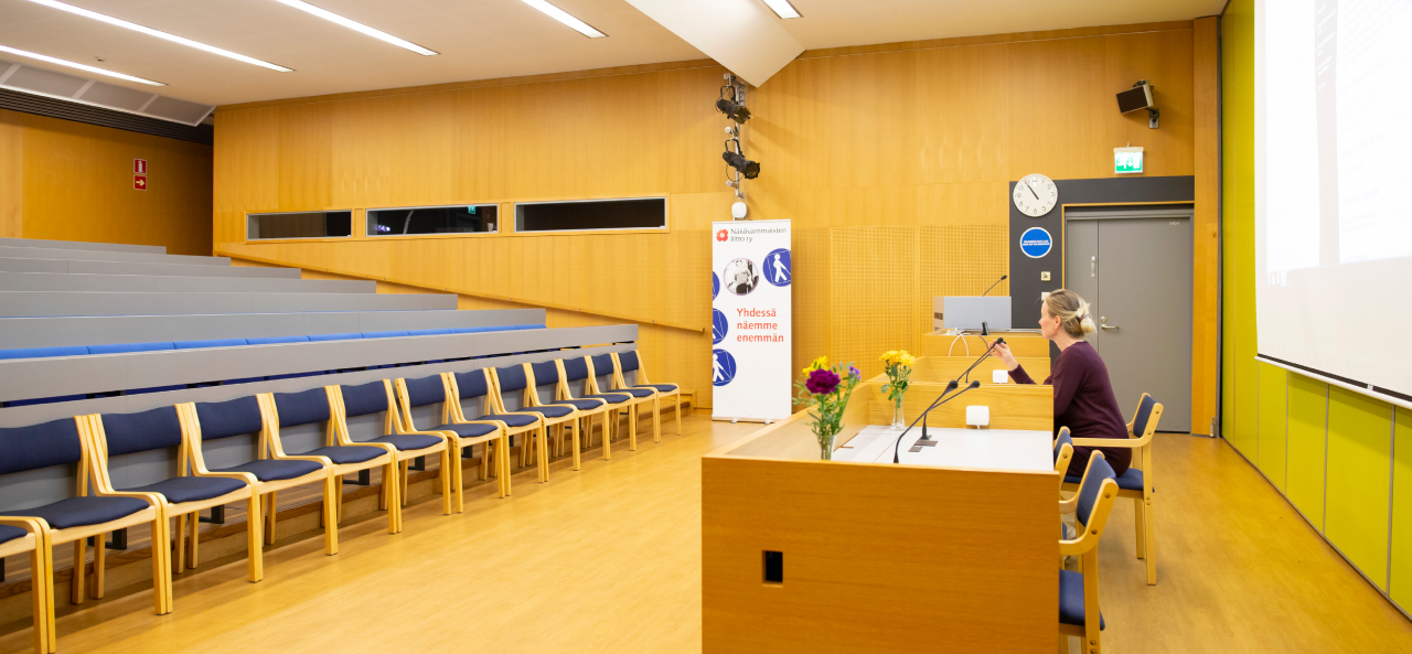 Nouseva auditorio, jossa harmaat penkkirivit. Edessä pyötä, jossa mikrofonit ja kukkakimppuja. Seinällä kello ja kaiuttimia, seinän edessä Näkövammaisten liiton roll-up.